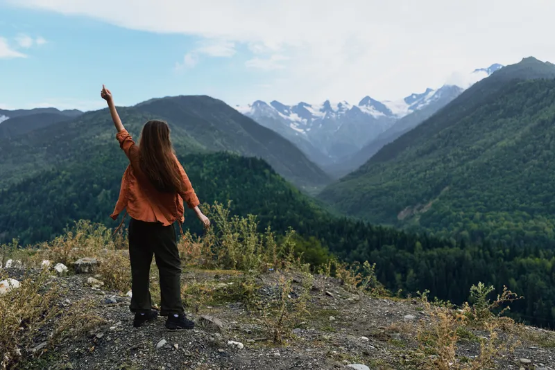 Female hiker stands on a rocky ridge overlooking a valley and snow capped peaks, lifting an arm in triumph, capturing a moment of adventure and vast natural beauty