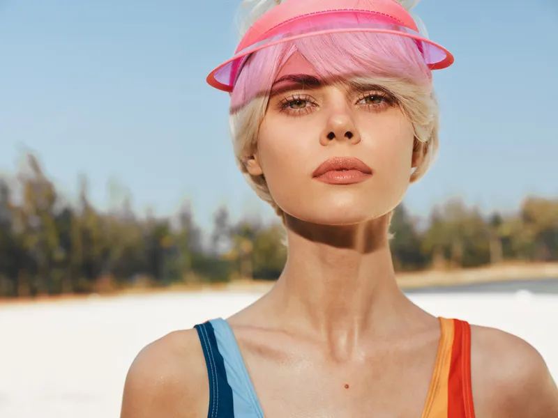 Close up portrait of a stylish woman at the beach wearing a pink visor, sunlit skin, short blonde hair, and colorful swimsuit straps, with a calm, confident gaze.