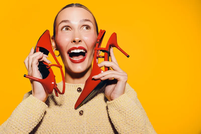 Happy woman in beige sweater holds red high heels near face, smiling widely on bright yellow background. Concept of fashion, joy, shopping, and vibrant lifestyle with colorful contrast.