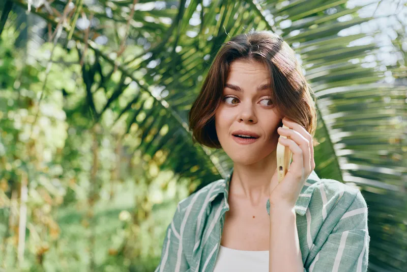 A young woman speaks on her smartphone outdoors, surrounded by lush foliage and sunlight, creating a fresh, natural portrait.
