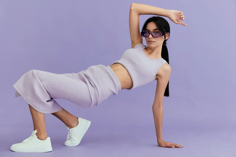 Young woman posing in casual purple outfit and white sneakers on lavender background, wearing sunglasses and showcasing modern street style with confident attitude.