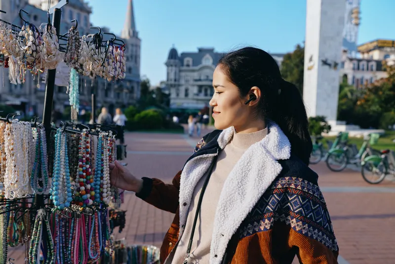Beads and jewelry stall outdoors, woman browses colorful accessories in a market setting, urban square in background, coat and scarf, calm shopper examining hanging strands