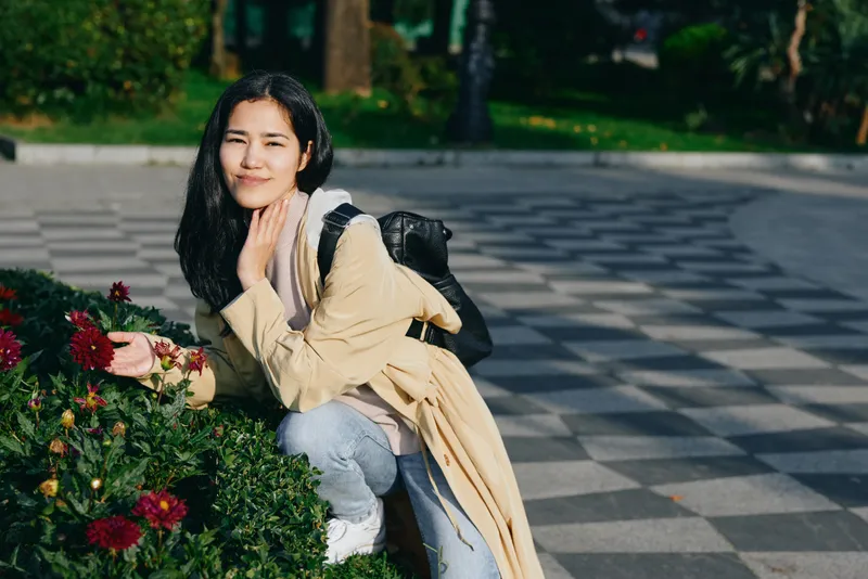 Woman in light coat kneels beside blooming flowers in a park, backpack on shoulder, enjoying a sunny outdoor moment with casual style and calm, reflective mood