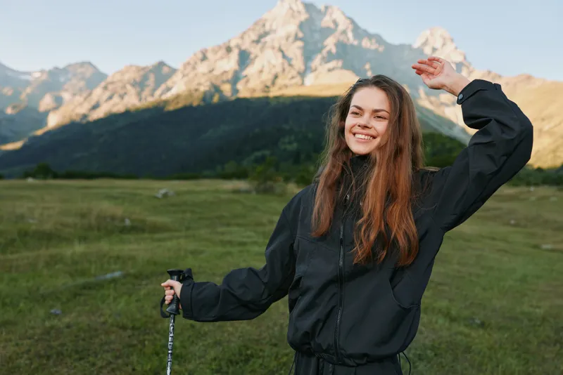 A cheerful young woman stands outdoors in a scenic mountain meadow, holding hiking poles and touching her hat, smiling warmly, capturing a bright, adventurous mood.