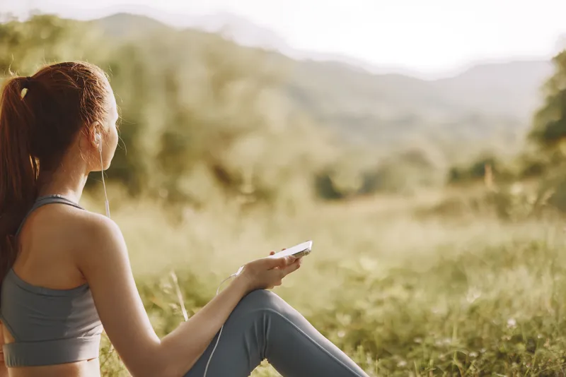 Young woman enjoying music in nature, wearing sportswear, sitting on the grass with a peaceful expression, ideal for wellness and fitness themes