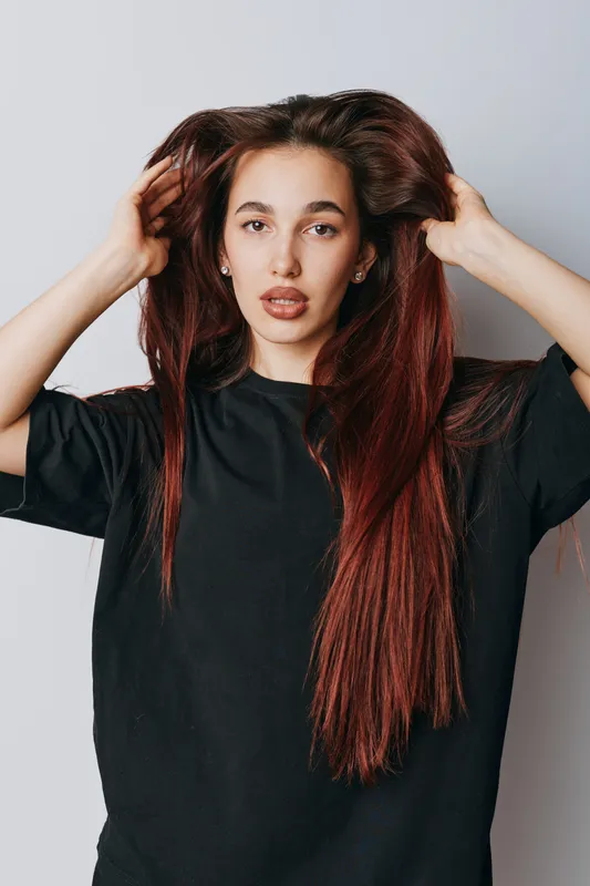 Young woman with long straight hair wearing black t shirt posing with hands in hair against white background, looking at camera with natural expression and minimal makeup.