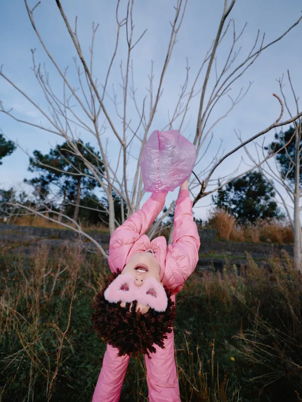 Child hanging from a bare tree in a pink coat, smiling with playful energy in an open field, wintery landscape under a clear sky, capturing a carefree outdoor moment.