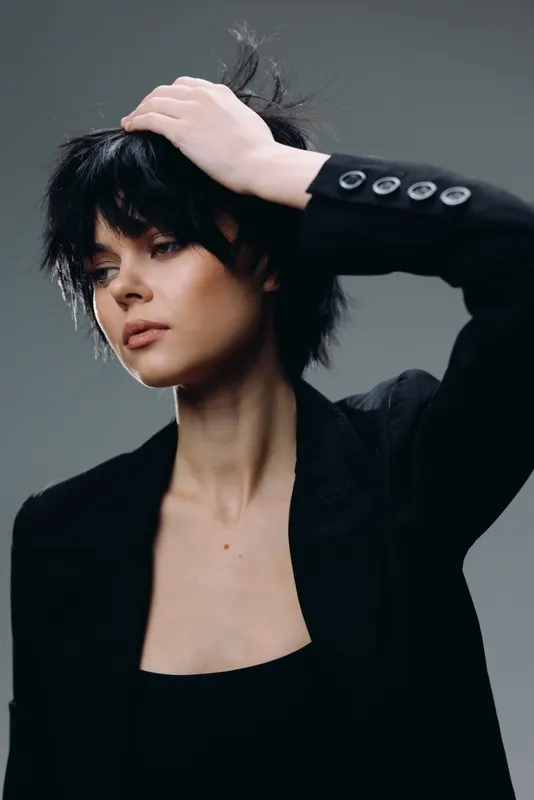 Stylish woman with short black hair wearing a black blazer posing with a thoughtful expression in a minimalistic studio setting.