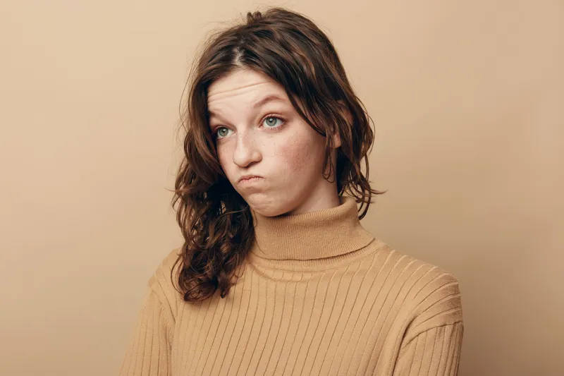 Young woman with curly brown hair wearing beige sweater looks confused and skeptical against plain beige background. Concept of doubt, uncertainty, and thoughtful expression.
