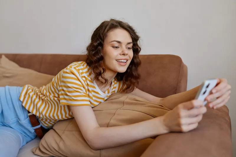 Casual woman lifestyle at home indoors, relaxed on a couch, smiling while taking a selfie, cozy atmosphere, soft light, natural colors, comfortable setting