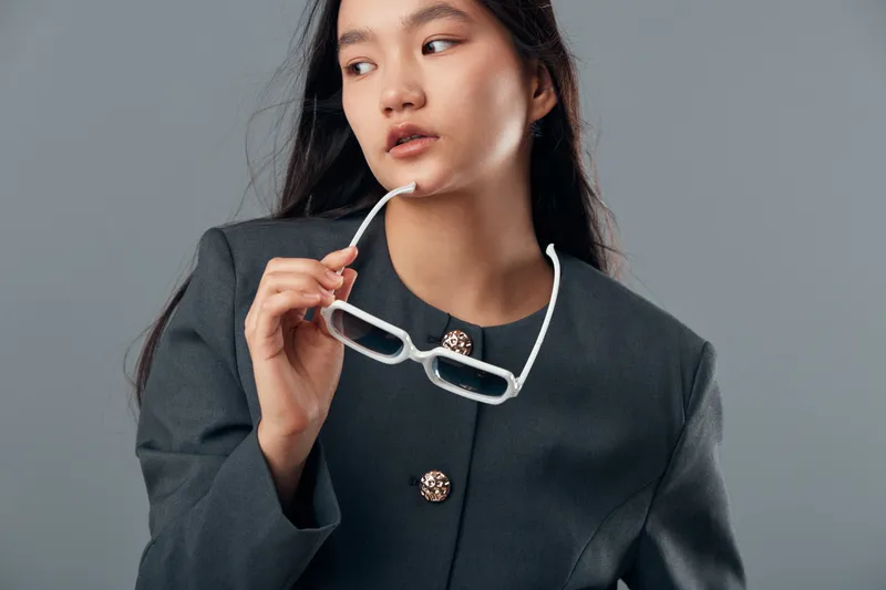 Woman with long dark hair wearing dark jacket holding white eyeglasses near face looking sideways on gray background. Female model posing thoughtful for fashion portrait with casual style.