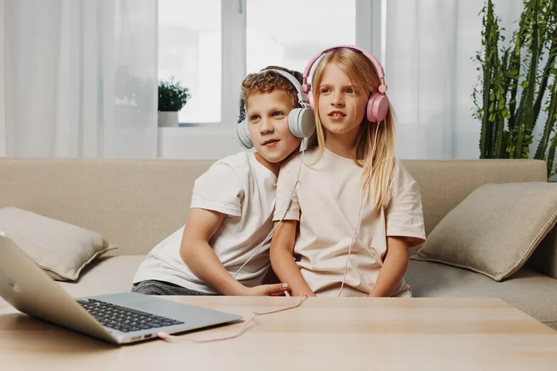 Two kids with headphones listen to music on laptop sitting on couch in cozy living room, enjoying leisure time together at home.