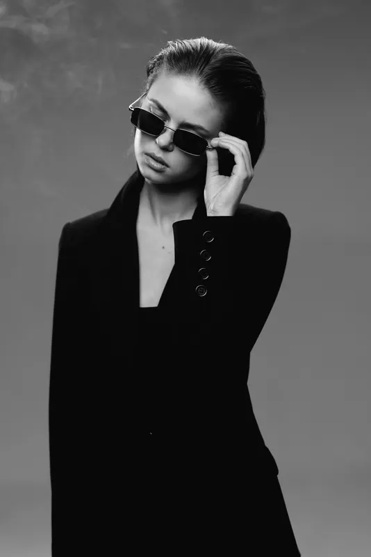 Black and white portrait of stylish woman wearing sunglasses and a blazer, expressing confidence and elegance.