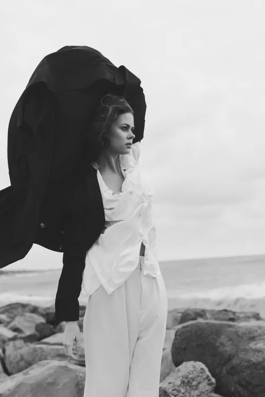 Woman in flowing coat on a rocky shore, wind catching fabric, monochrome coastal scene with dramatic fashion mood, sea spray and moody sky.