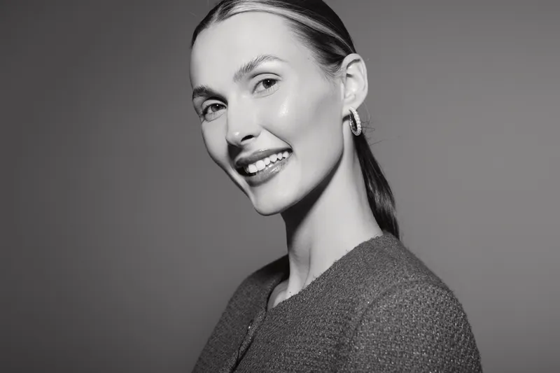 Smiling white woman with light skin and pulled back hair in sweater poses in studio. Black and white portrait with soft lighting creates a calm and elegant mood.