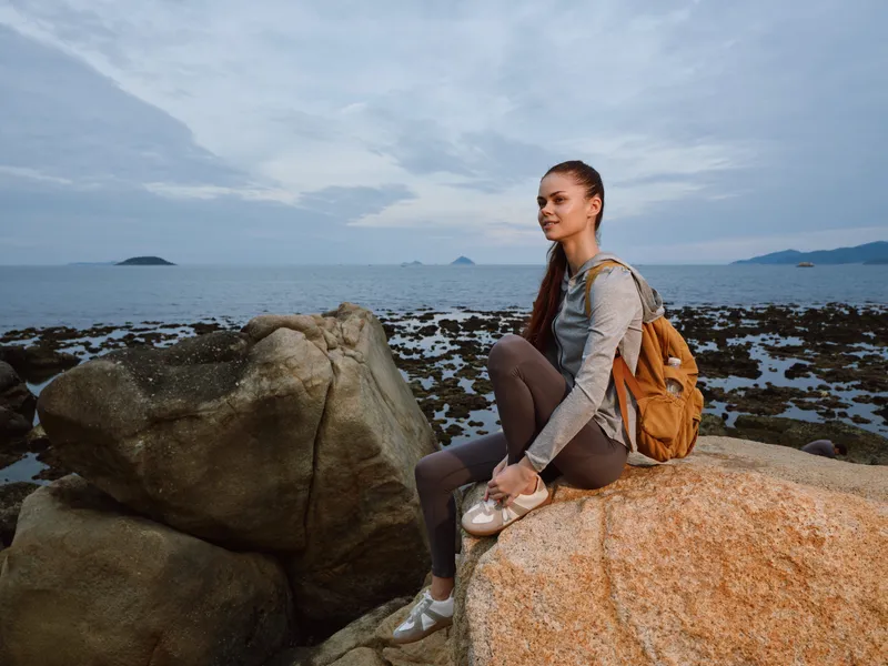 Young woman with a backpack sitting on a rock by the seashore, displaying a calm expression, wearing casual clothing suitable for an outdoor adventure