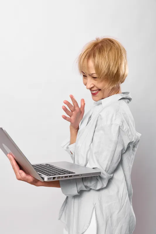 Smiling woman doctor waves toward camera while working on a laptop in a white coat, plain background, conveying healthcare communication for remote support.