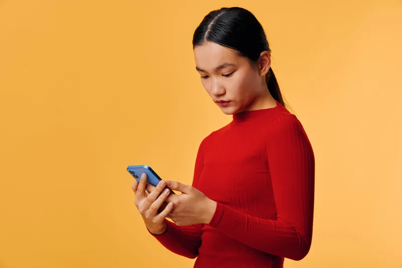 Woman with black hair and red sweater using smartphone against orange background for communication messaging and technology concept in casual style.