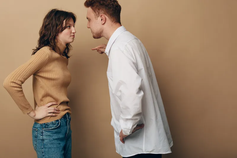 A young man and woman argue facing each other with intense expressions. The woman stands with hands on hips, the man points finger, both in casual clothing on beige background.