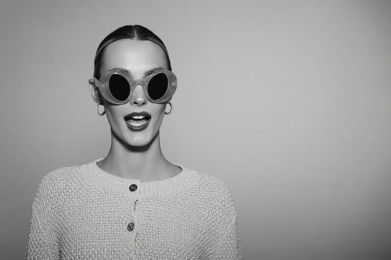 Young Caucasian woman with light skin wearing round sunglasses and light sweater expresses surprise or excitement in studio.