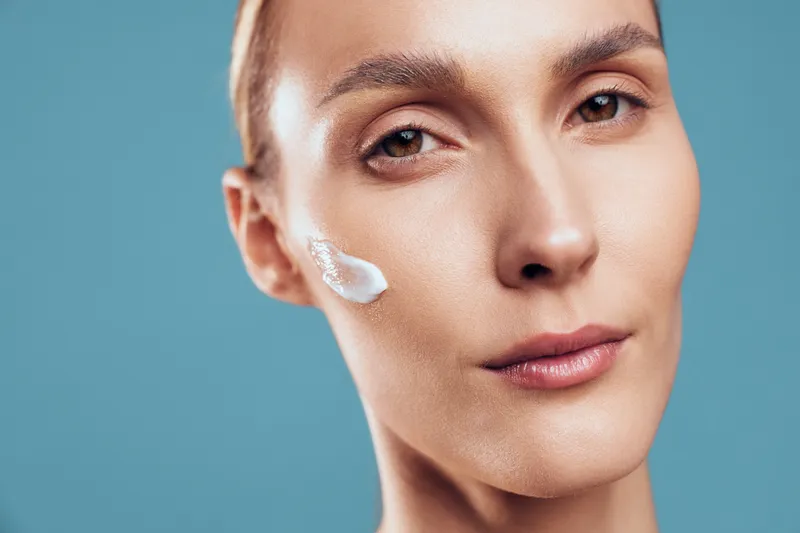 Young woman with brunette hair applies white face cream on her cheek in a close up portrait with a blue studio background, conveying a precise skincare moment