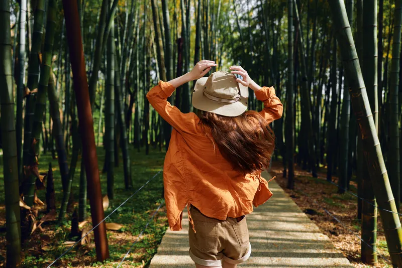 Woman hat bamboo forest pathway walking orange back in casual straw hat and loose shirt, summer travel and nature scene on wooden trail with sunlight, greenery and tall stems.