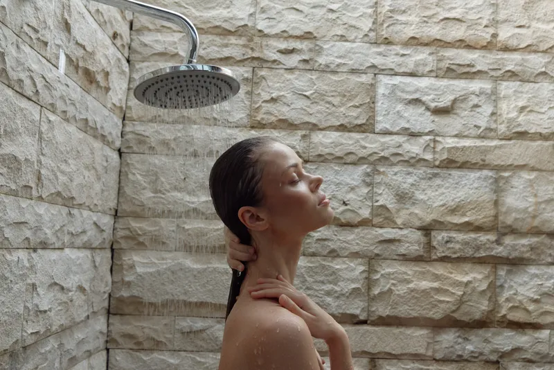 Woman enjoying relaxing shower under rain showerhead with closed eyes, wet hair, and natural stone wall background in serene bathroom setting.