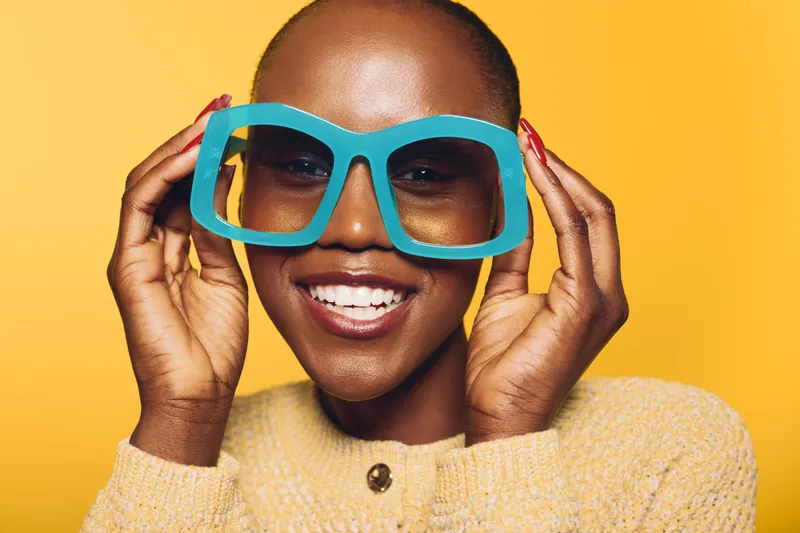 Smiling black woman with dark skin wearing large blue sunglasses and beige sweater poses against yellow background. Concept of fashion, confidence, beauty, and modern style portrait.