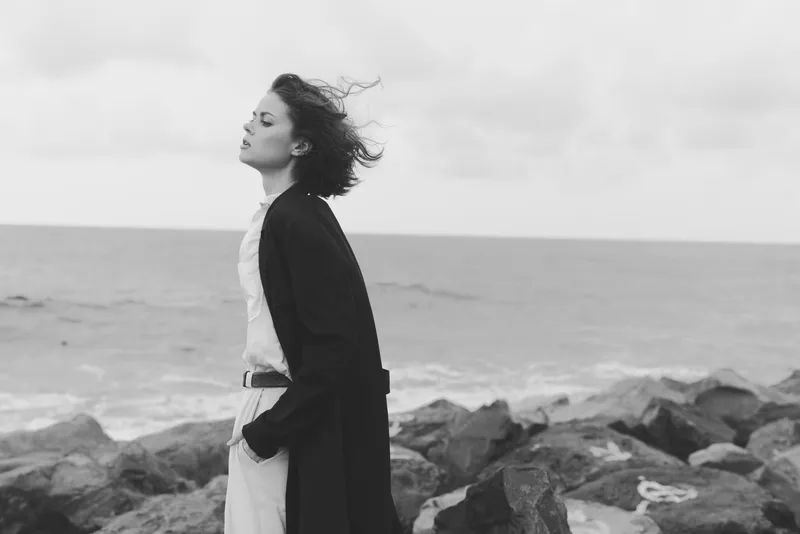 Woman stands on a rocky coast facing the sea, wind in hair, wearing a long coat over light attire, monochrome scene conveying solitude, resilience, and reflective mood by the shoreline