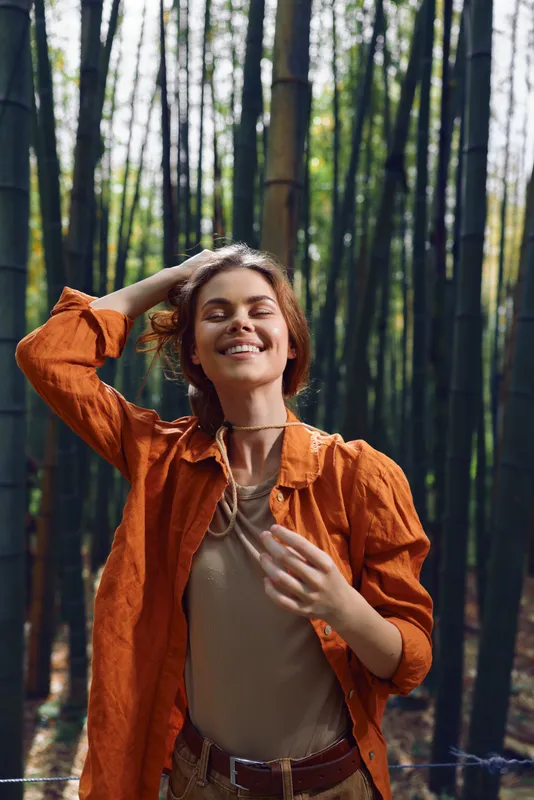 Woman smile bamboo forest orange portrait nature happiness relaxed outdoors, smiling with eyes closed wearing orange shirt and layered necklace, peaceful casual lifestyle in green grove with sunlight