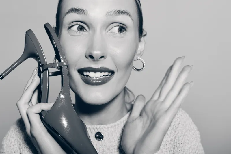Young white woman with light skin holds high heel shoe near face smiling, looking sideways. Studio portrait in black and white with joyful expression, fashion and beauty concept.