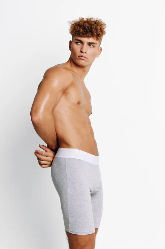 Young man with curly hair posing shirtless in gray shorts against white background. Standing side profile, looking to the side with hands behind back showing casual fitness and body posture.