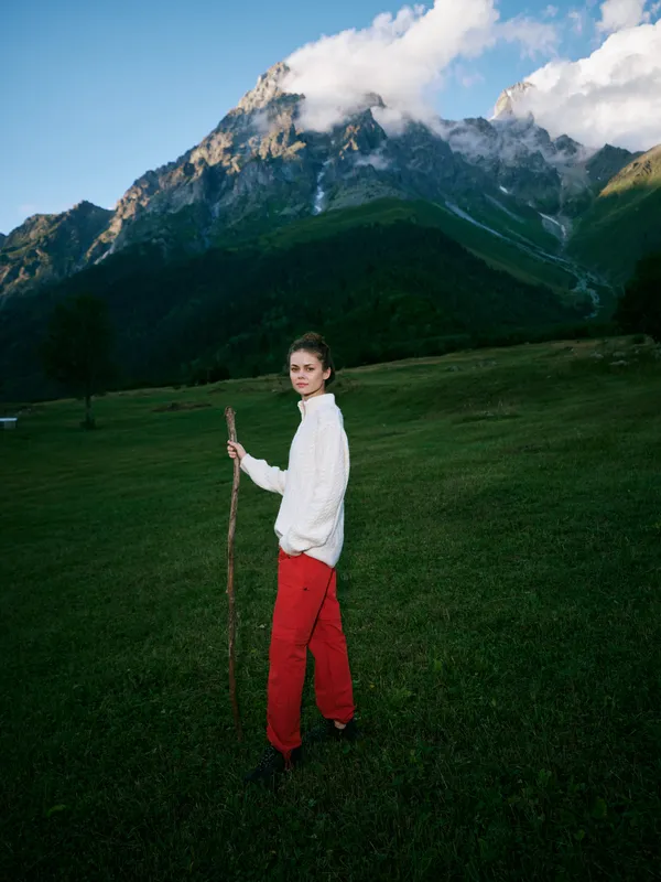Hiker in white sweater and red pants stands in a wide meadow, mountains looming behind, gripping a wooden staff, ready for exploration and outdoor adventure under blue sky.
