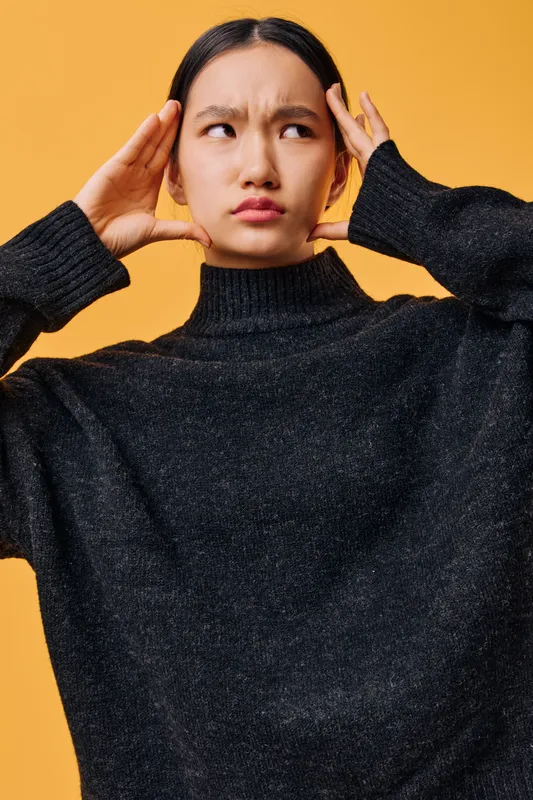 Woman with black hair and black sweater holding her head showing confused and frustrated expression on orange background. Portrait of young Asian female with thoughtful look.