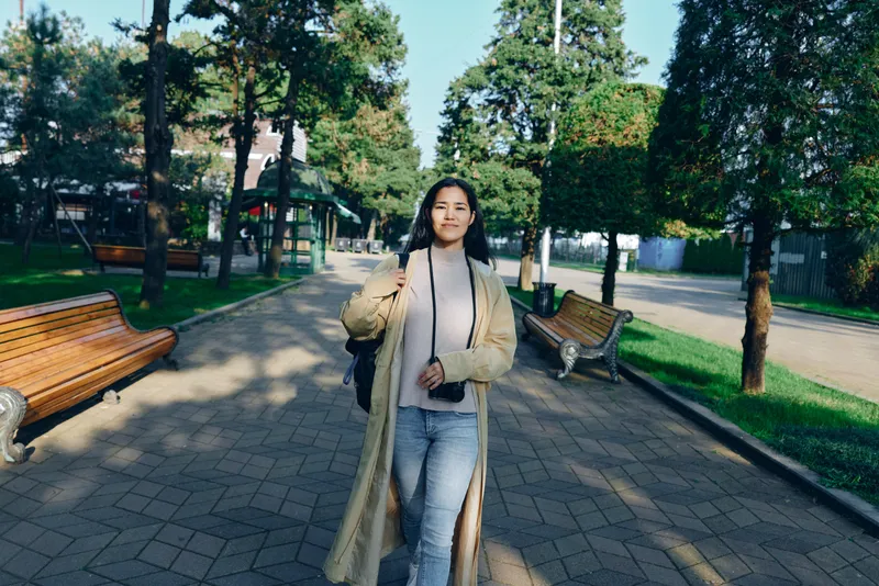 Woman wearing a long beige coat walks along a tree lined park path, carrying a bag, casual stylish urban stroll, sunlight filtering through leaves, serene morning atmosphere, candid lifestyle shoot