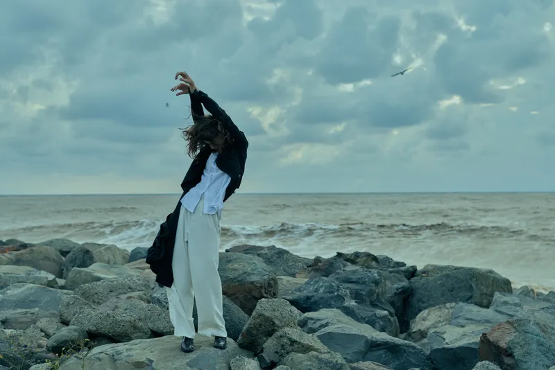 Woman on rocks by the sea, movement captured with wind sweeping clothing, outdoor scene along the coast, dress and jacket framing a dramatic, moody moment.