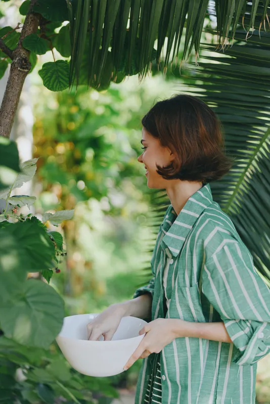 woman in a striped green robe stands outdoors in a lush garden, holding a white bowl, gentle smile, serene mood, natural light, peaceful moment, candid portrait, nature