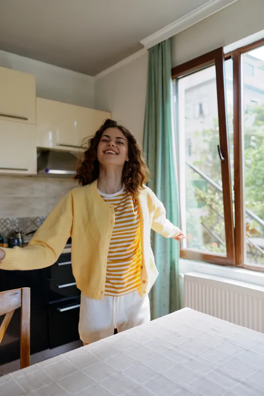 Cheerful woman indoors, standing in a bright kitchen, arms open, wearing a yellow cardigan and striped top, enjoying a sunlit moment, relaxed mood, soft natural light, casual home scene, lifestyle