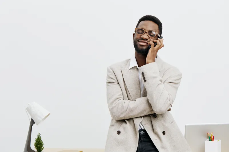 Confident young African American man wearing beige blazer and glasses smiling thoughtfully while talking on mobile phone in bright modern office setting. Studio portrait of professional person.