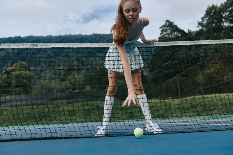 Young female tennis player in a stylish sports outfit reaching for a tennis ball on a court surrounded by a lush green landscape, showcasing athletic energy and determination