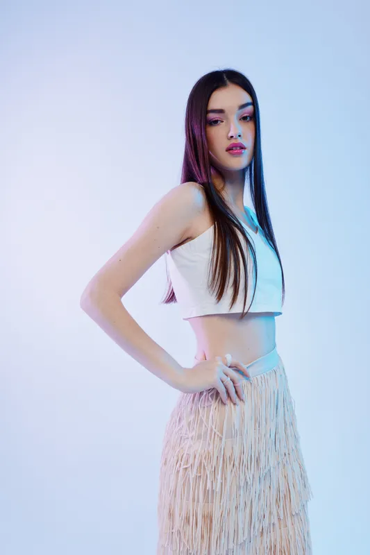 Young woman with long dark hair poses confidently in a white crop top and fringed skirt. Studio shot with soft blue lighting. Concept of beauty, fashion, and style.