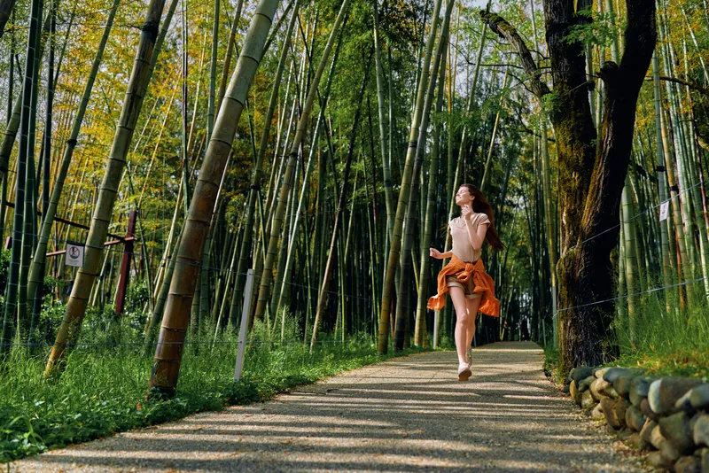 Woman bamboo forest path running in orange skirt with sunlight dappled through canopy, joyful motion on a shaded trail among green trees and foliage, serene outdoor nature scene.