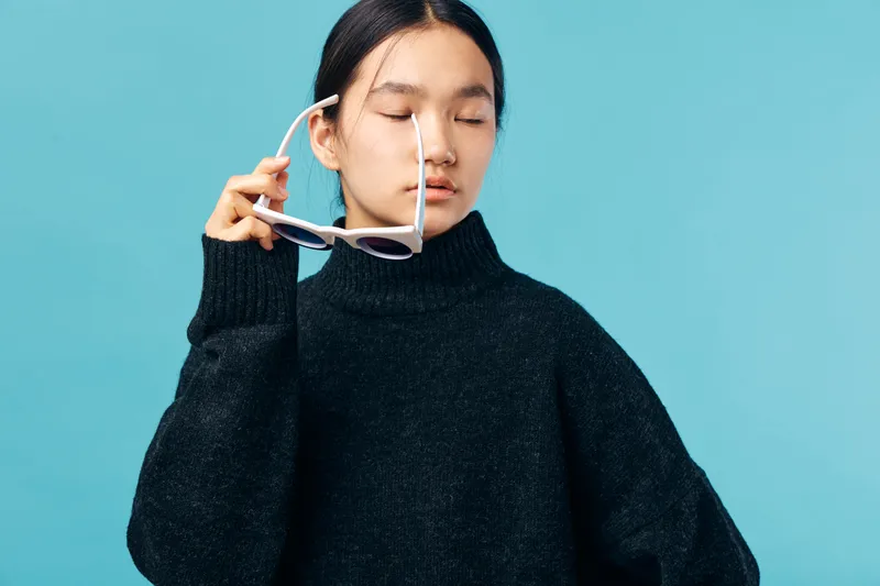 Young woman in black sweater holding sunglasses with eyes closed studio portrait on blue background. Female model with calm expression and casual style for fashion or lifestyle concept.