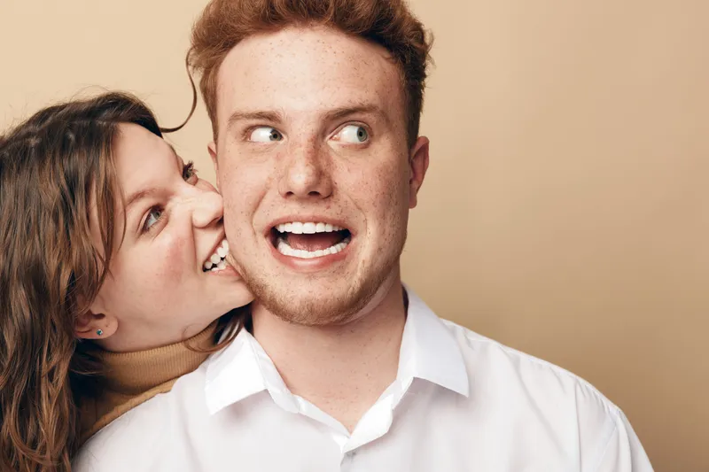Playful young woman whispers in surprised young man’s ear. Both have casual clothes and light background. Concept of friendship, fun, communication, and joyful emotions.