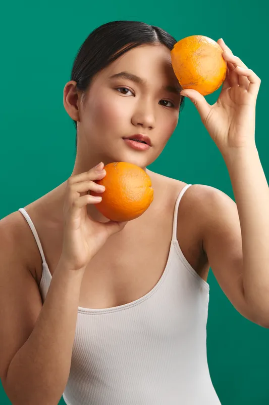 Asian woman holds oranges against a vivid green backdrop, posing with a soft smile and relaxed pose. The scene conveys freshness, health, and a playful yet calm mood.