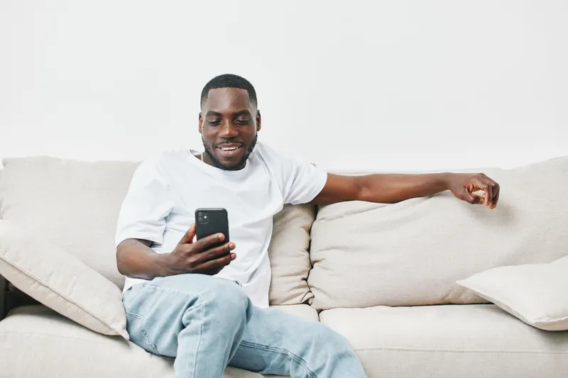 Smiling Black man relaxing on a sofa, using a smartphone, casually dressed in a white t shirt and jeans, exuding a joyful and content vibe in a modern living room
