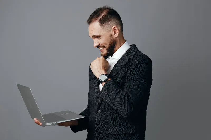 A professional businessman holds a laptop against an isolated colored background, demonstrating confident mood, focus, and subtle pride during a moment of achievement.