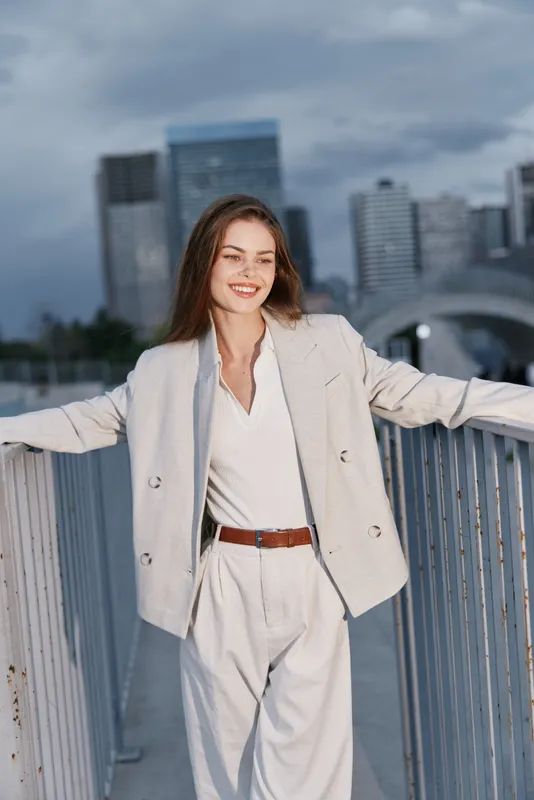 Smiling woman in light business suit standing outdoors with city skyline background, confident young Caucasian professional in beige coat and trousers
