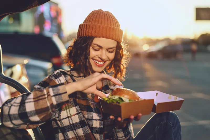 Woman, burger, takeout, smile, outdoor, car, beanie, parking: Young woman smiles over a takeout burger by her parked car at golden hour, candid lifestyle shot with authenticity and emotional