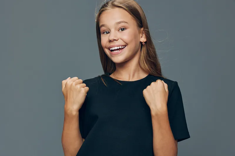 Happy girl smiling and showing fists with excitement and joy on gray background. Portrait of young child with positive expression and casual black t shirt for success.